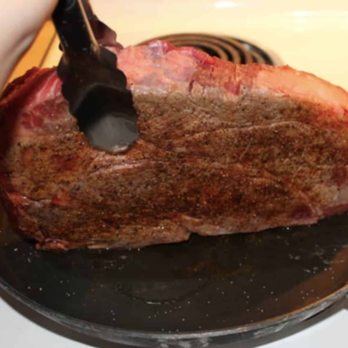 tongs holding a chuck roast in a frying pan so it can sear all the edges
