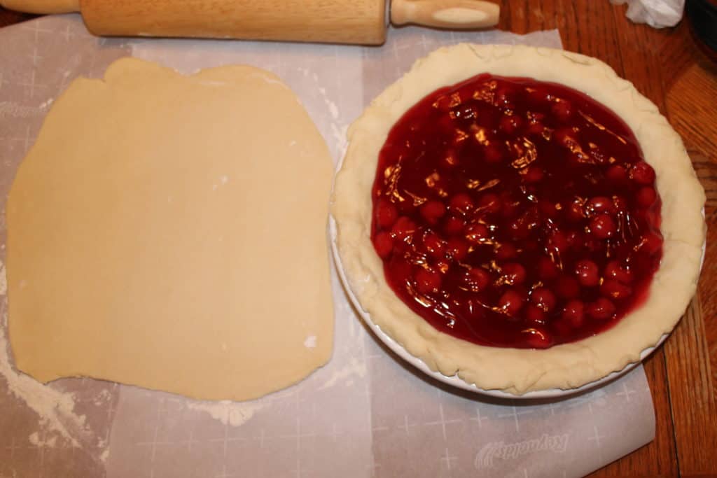 cherry pie filling added to the ceramic pie dish with the dough. Another pie dough rolled out sitting beside it to be used for the top of this cherry pie