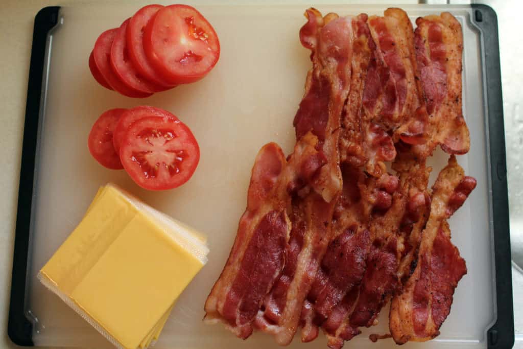 Cutting board with cooked bacon, sliced tomatoes, and slices of cheese.