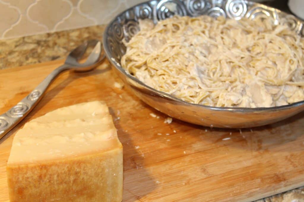 a large silver serving bowl with chicken alfredo next to the bloc of parmesan cheese