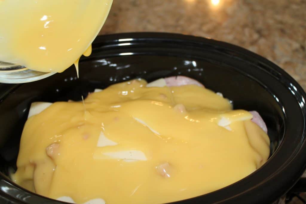 Pouring chicken gravy into the crock pot with chicken, cheese, and ham.