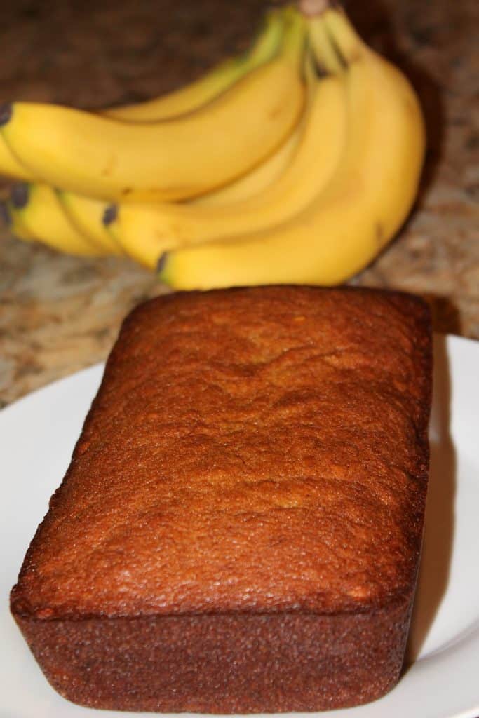 Banana bread loaf on a white plate with bananas behind it