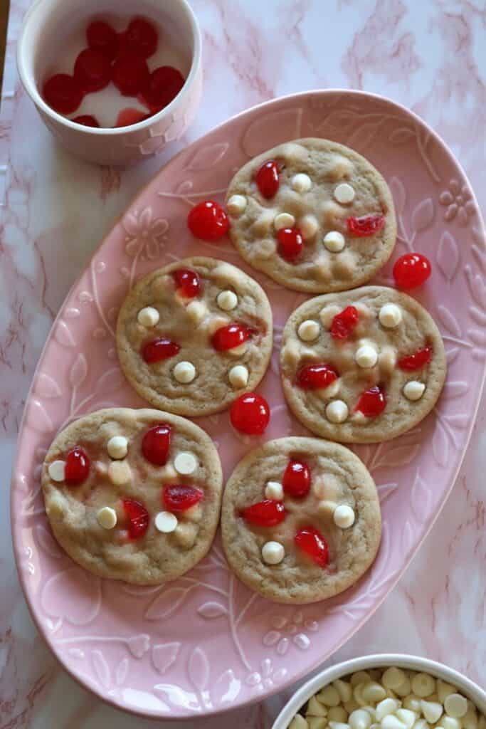 a plate with cherry white chocolate chip cookies and two small bowls with extra white chocolate chips and maraschino cherries